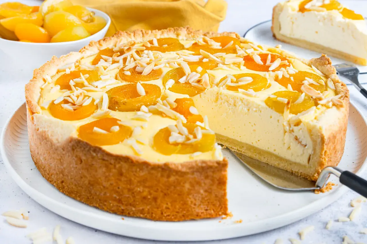 Ein köstlicher Landfrauen Aprikosenkuchen mit Mandelsplittern, frisch angeschnitten. Daneben eine Schüssel mit Aprikosen.