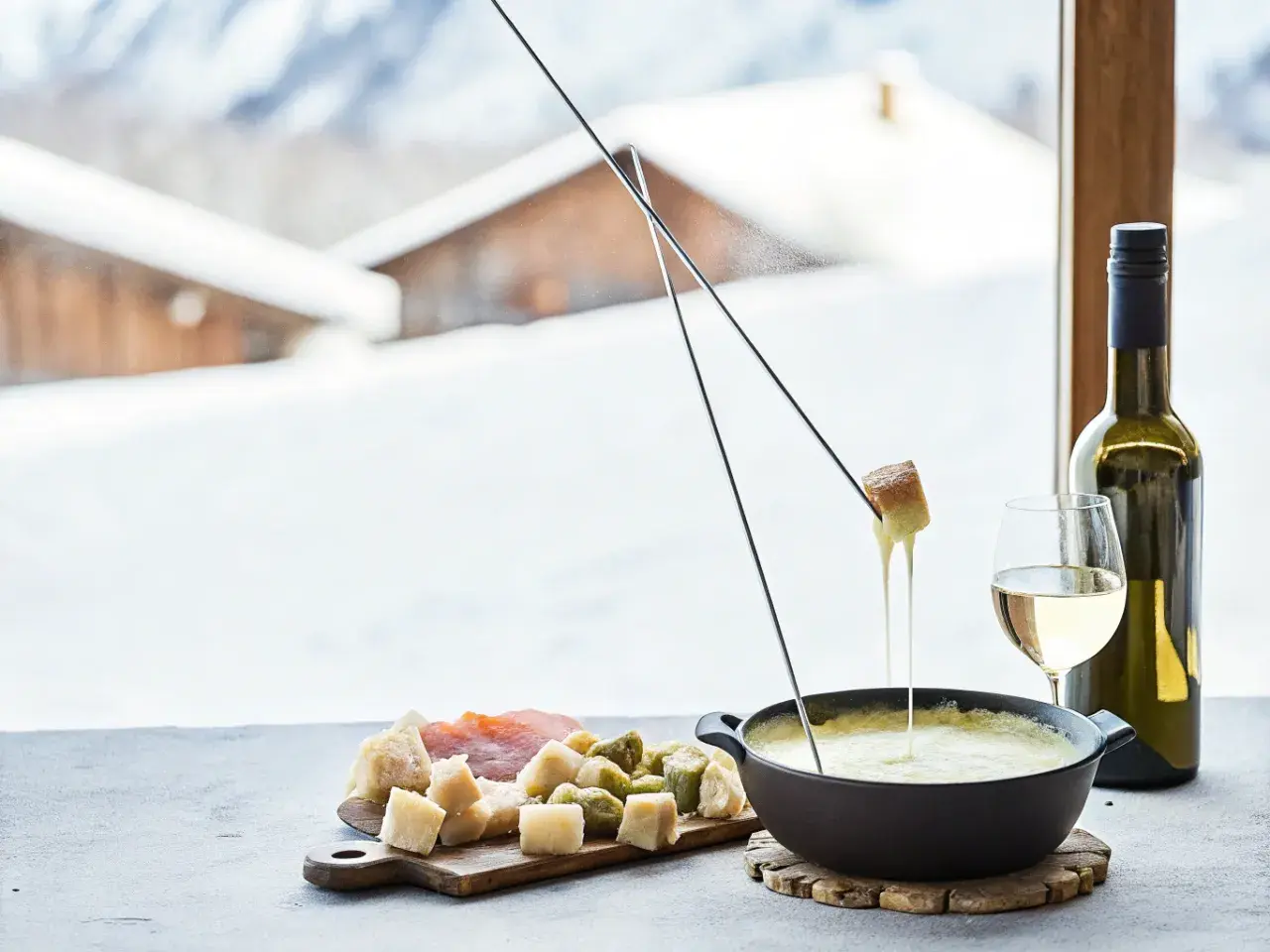Une délicieuse recette fondue au fromage, avec du pain et des cornichons, servie avec du vin blanc devant un paysage enneigé.