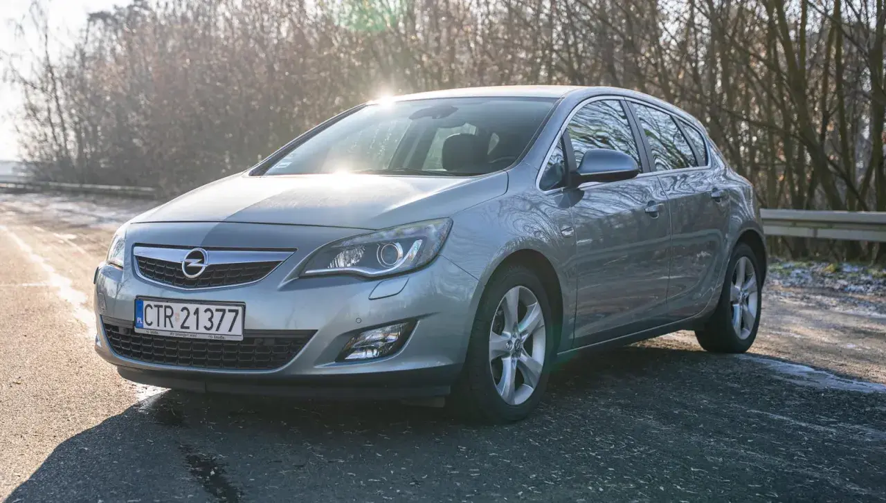 Szary Opel Astra 2011 na drodze, z drzewami w tle i słońcem odbijającym się od karoserii.