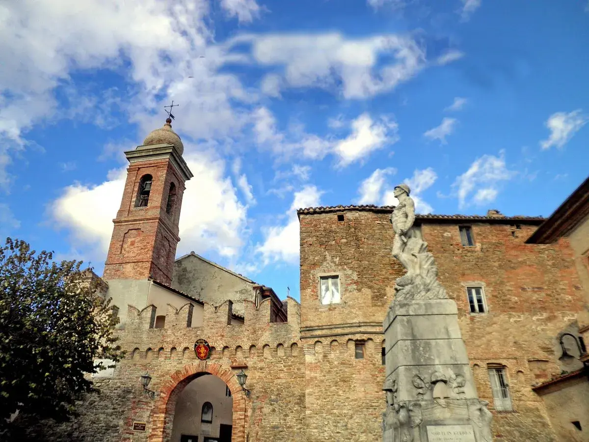 Poggio San Marcello: campanile in mattoni, un arco fortificato e una statua di bronzo sotto un cielo azzurro con nuvole bianche.