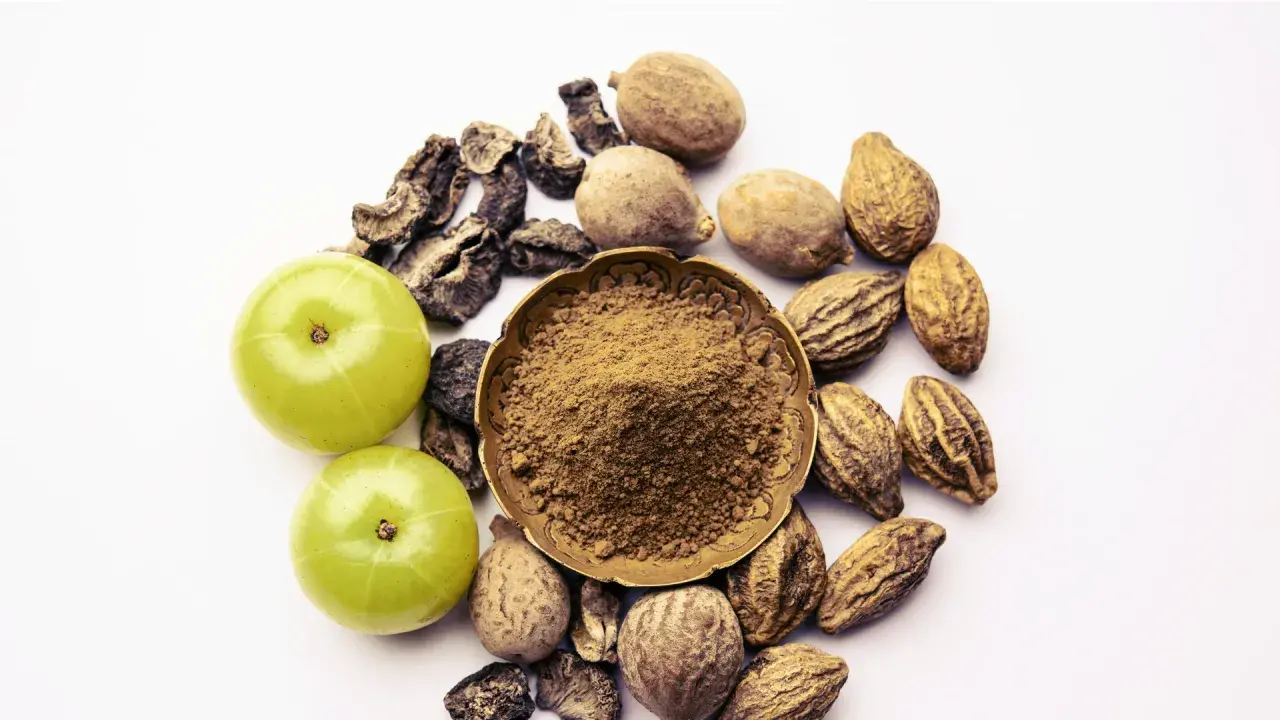 Triphala Pulver in einer Schale, umgeben von Amla-Früchten und getrockneten Haritaki- und Bibhitaki-Samen. Die Triphala Wirkung wird durch diese Zutaten erzielt.