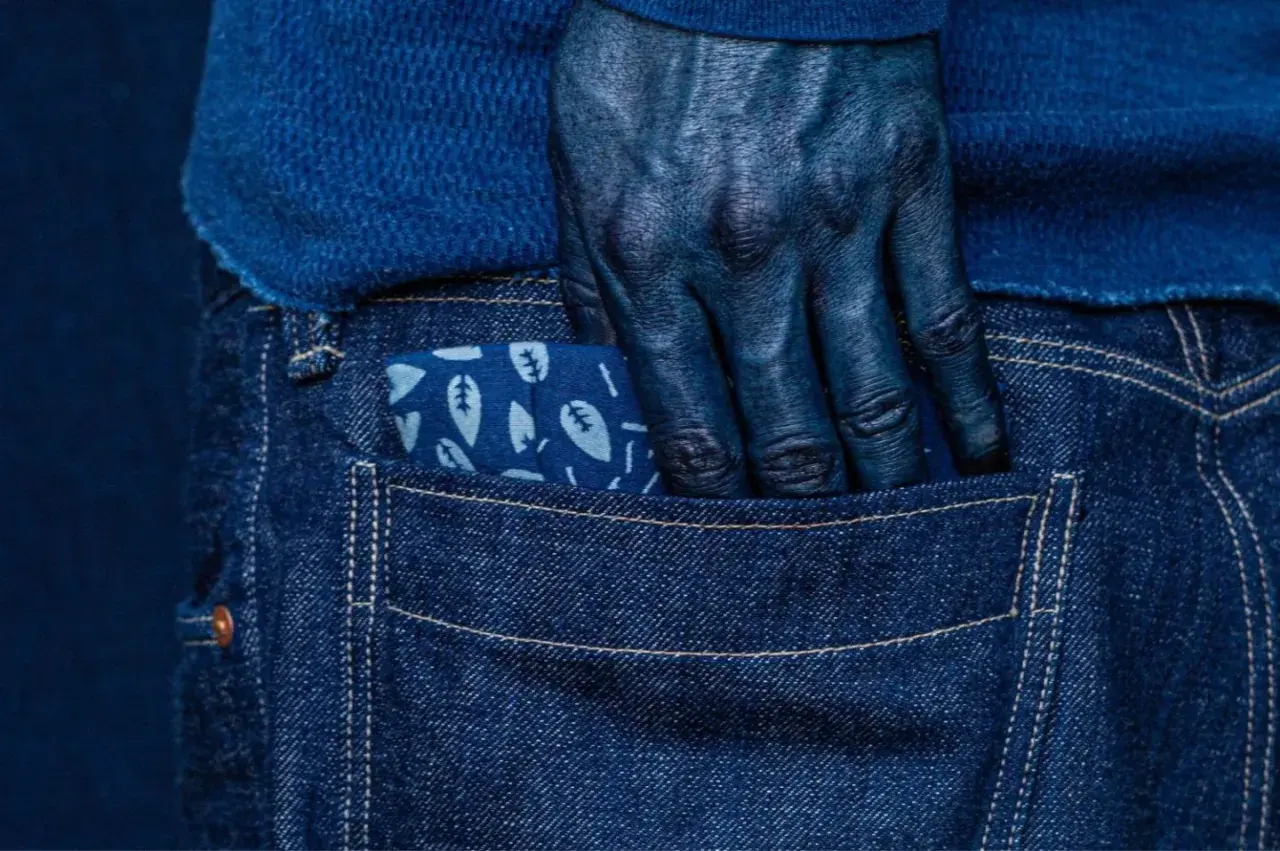 Main bleue, main plongée dans la poche d'un jean, le tissu orné de feuilles, témoignage de la teinture.