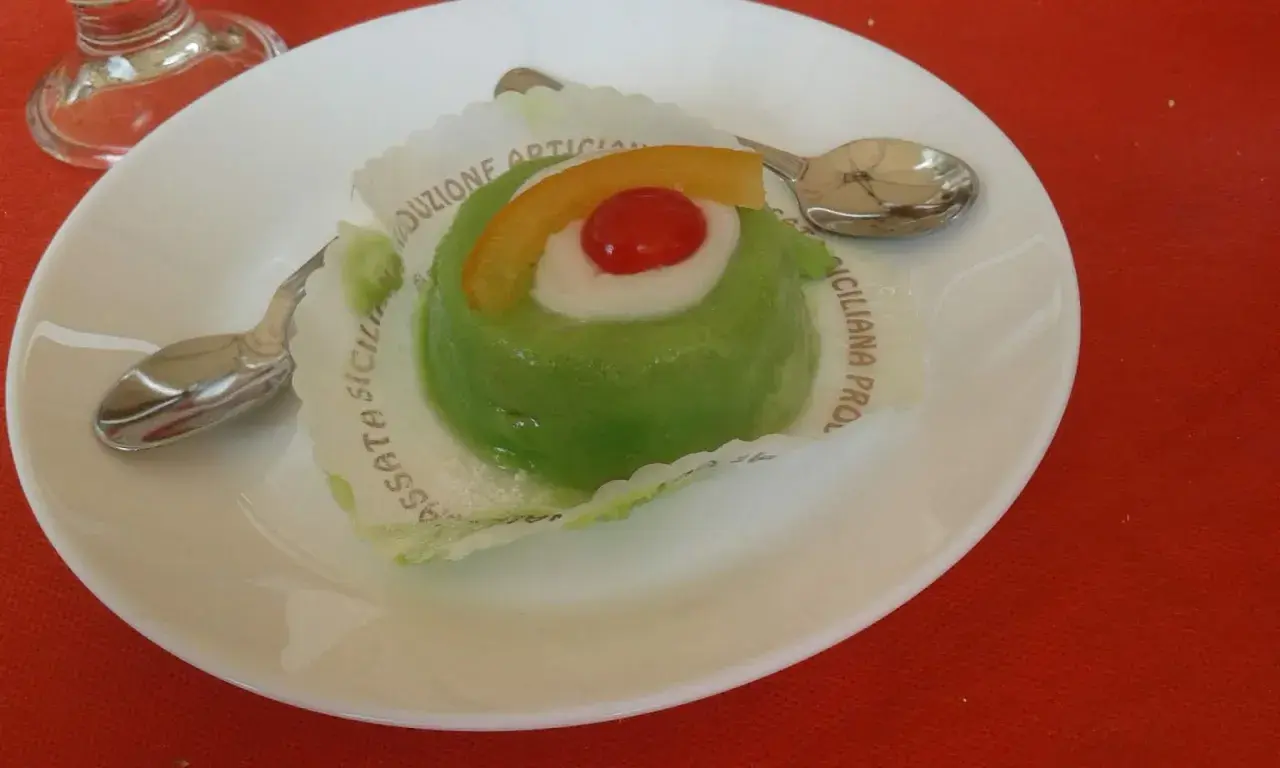 Zielone lody cassate, ozdobione wiśnią i skórką pomarańczy, na białym talerzyku z łyżeczką.