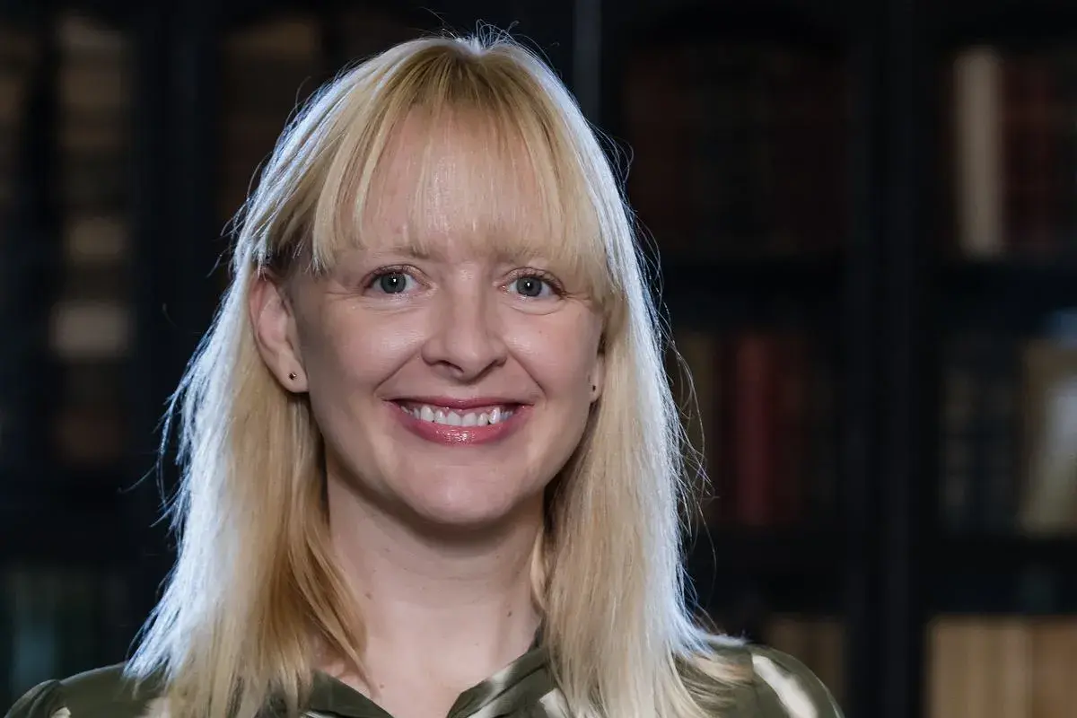 Uśmiechnięta blondynka z grzywką, w tle biblioteka. Jej spojrzenie sugeruje, jakie są relacje między autorem a jego literackim alter ego – pełne refleksji.