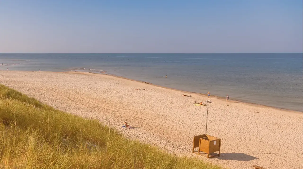 Słoneczna plaża w Kuźnicy nad morzem. Kilka osób odpoczywa na piasku, w oddali widać spokojne morze i błękitne niebo.