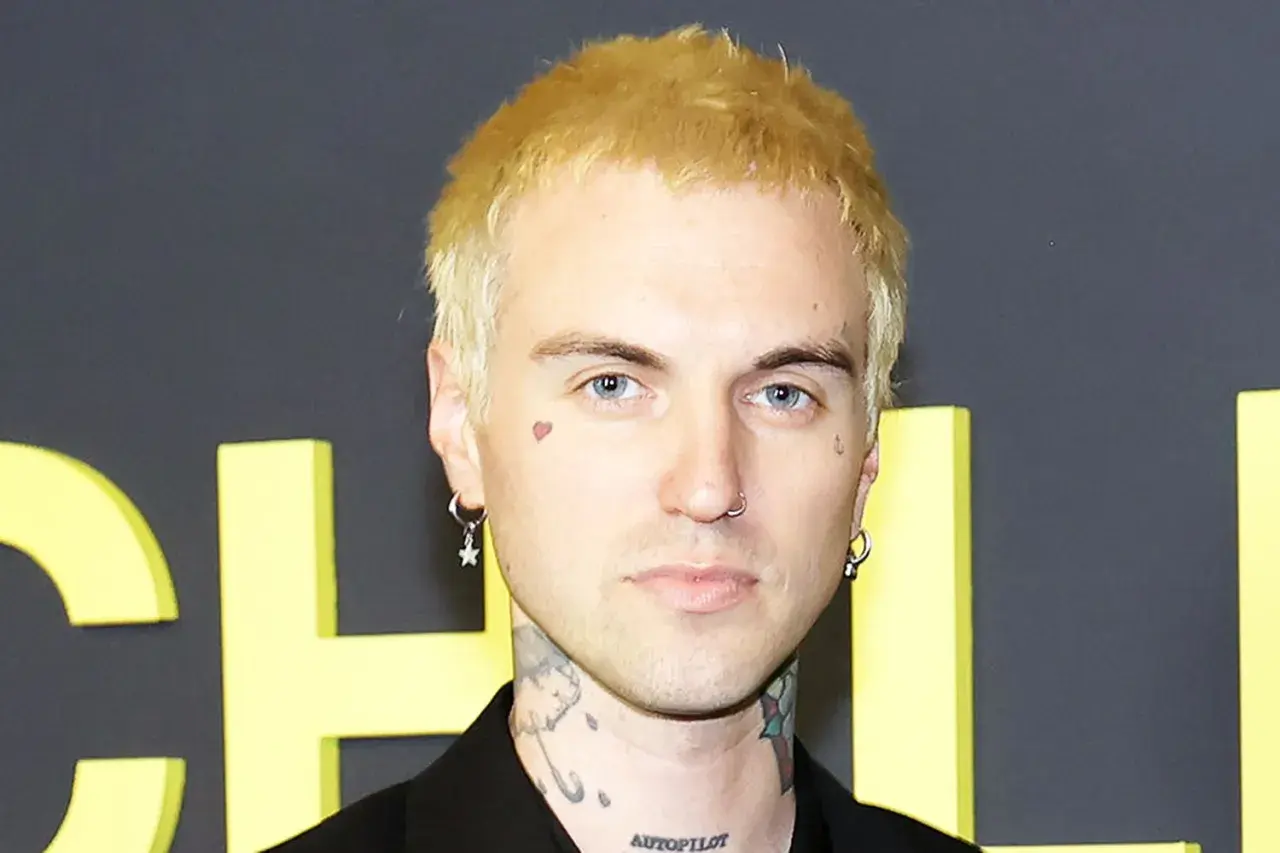 A young man with blonde hair, blue eyes, and tattoos, including "AUTOPILOT" on his neck, poses for a photo. He wears hoop earrings and a nose ring. His style is reminiscent of jordan jutes.
