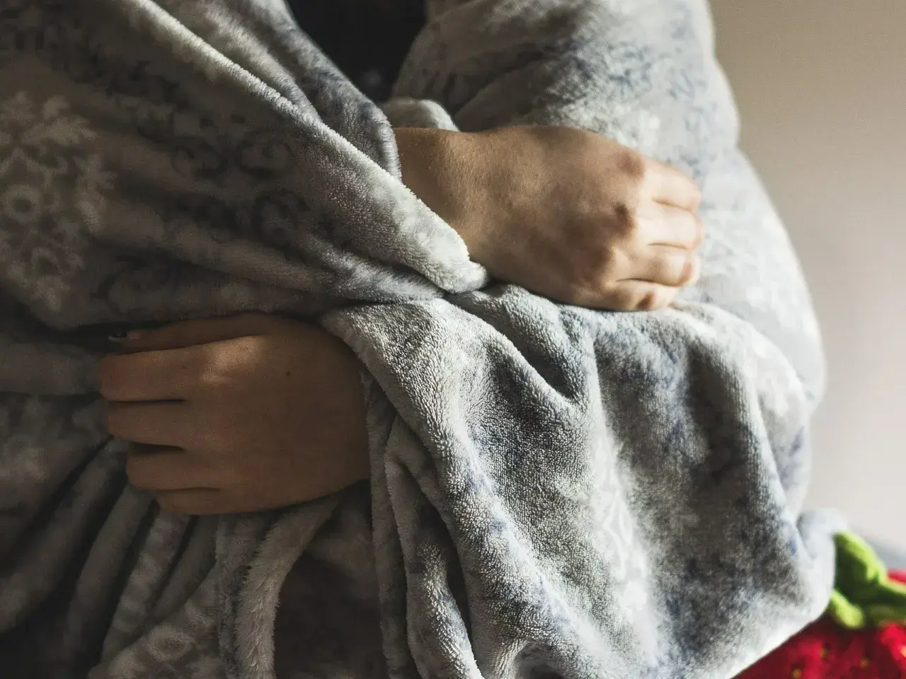 Dłonie skrzyżowane na piersi, otulone miękkim kocem, symbolizują ciągłe uczucie zimna i potrzebę ciepła.