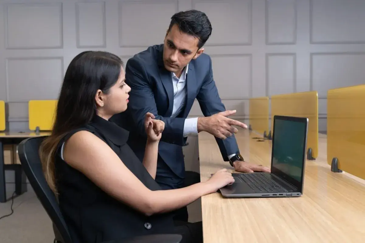 Un uomo in giacca e cravatta indica lo schermo di un laptop a una donna seduta. Potrebbe essere un responsabile risorse umane al lavoro, che discute un progetto.