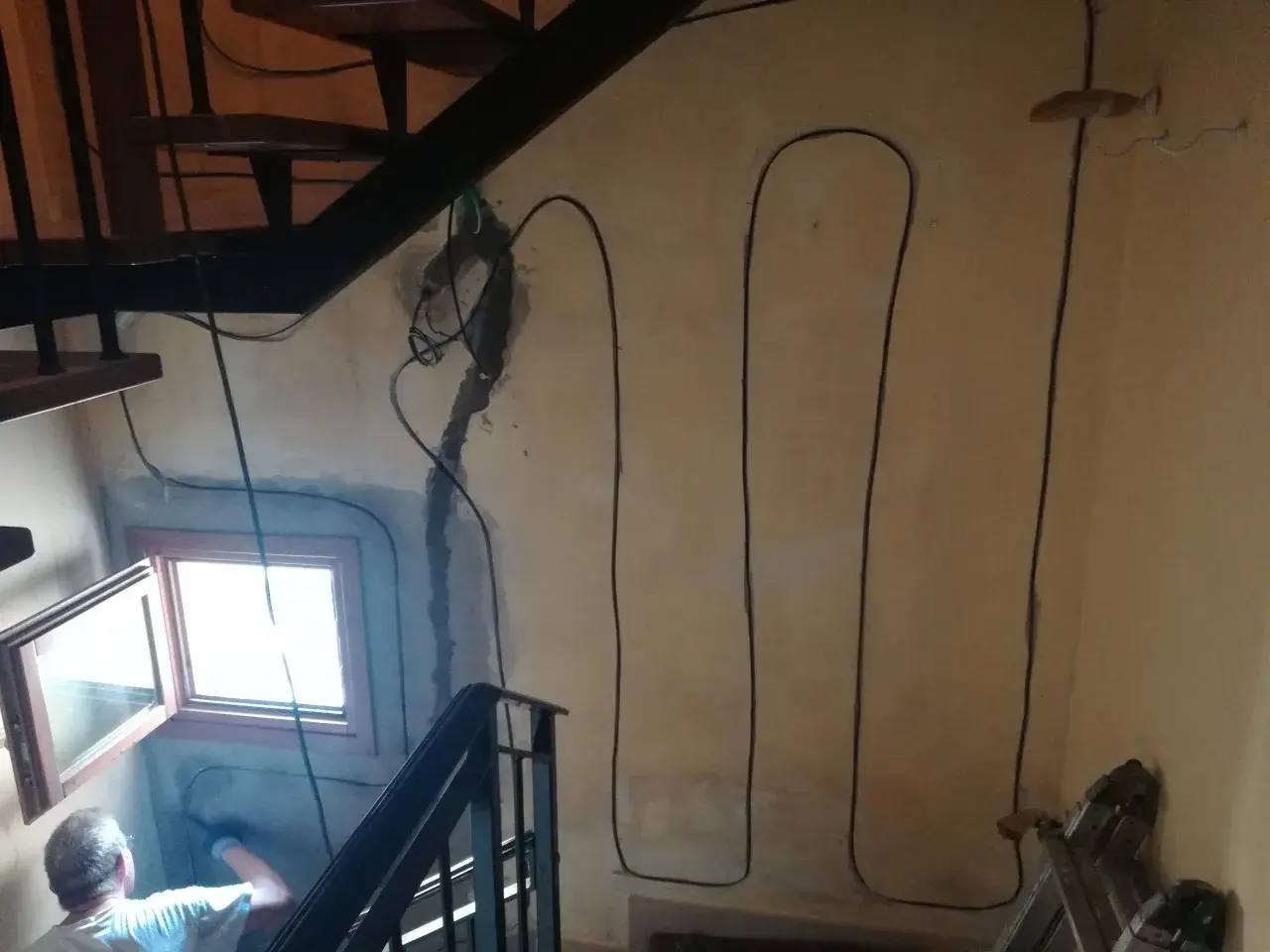 Lavori in corso per l'isolamento pilastri: cavi neri corrono lungo la parete accanto a una scala e una finestra.