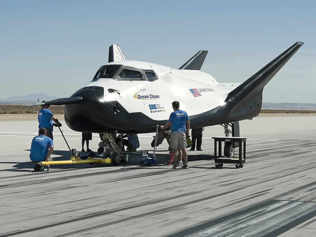 Dream Chaser, kosmiczny pojazd, przygotowywany do startu. Zespół techników pracuje przy jego podwoziu na pustynnym lotnisku.