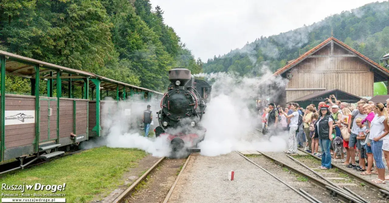 Para rodzinna podróż w Bieszczady z dziećmi. Parowóz z wagonami czeka na pasażerów, otoczony parą i zielenią.