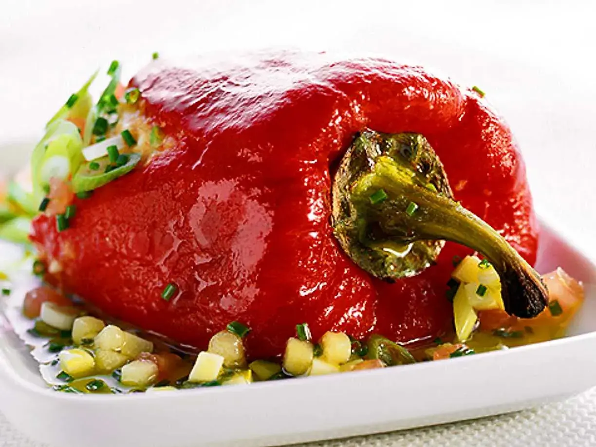 En fylld, röd paprika serveras på en vit tallrik med en sallad av tärnad paprika och örter. Ett läckert recept med grillad paprika på burk.