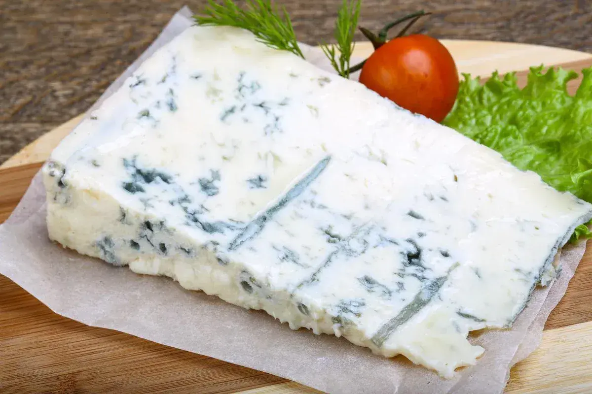 Un pezzo di gorgonzola formaggio con venature blu, guarnito con pomodorino e lattuga su tagliere di legno.