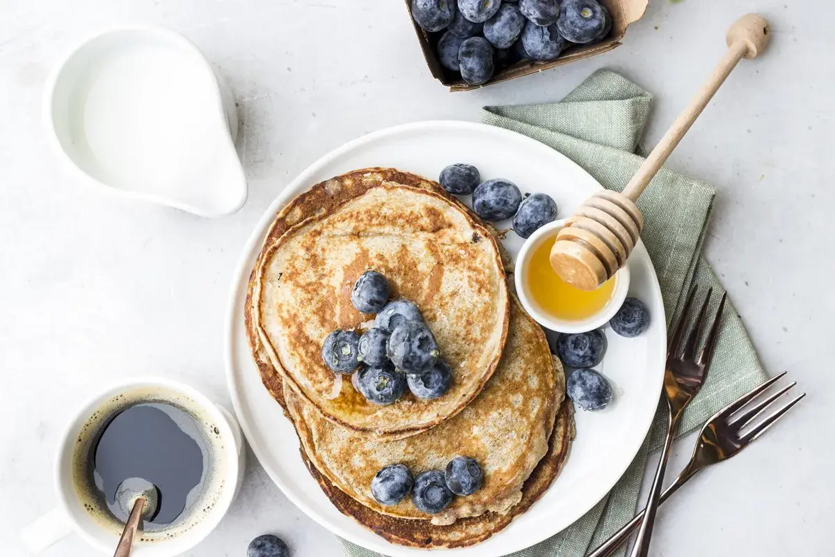 Pancake senza uova con yogurt, guarniti con mirtilli freschi e miele. Un caffè nero e una brocca di latte completano questa deliziosa colazione.