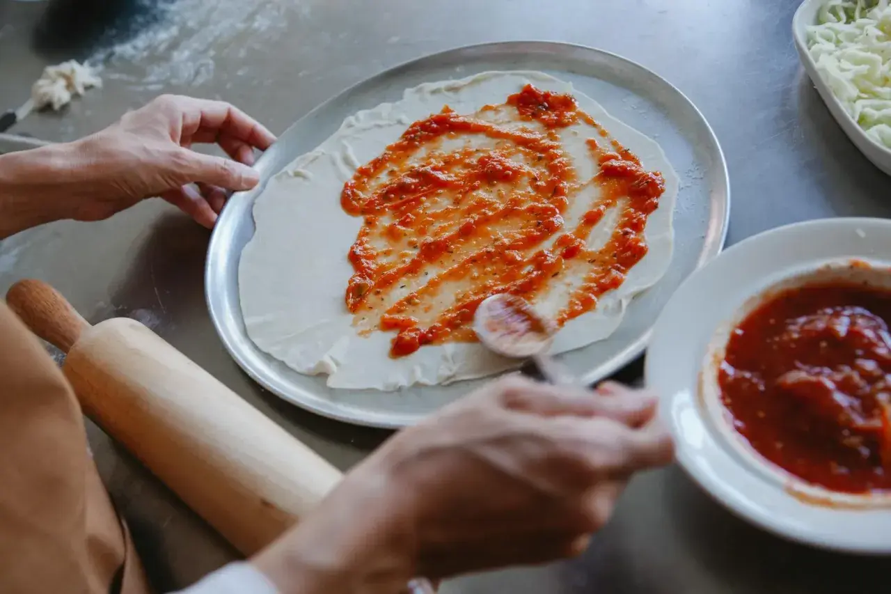 Ręce rozsmarowują pyszny sos pomidorowy do pizzy na cieście. Obok miseczka z sosem i tarta mozzarella.
