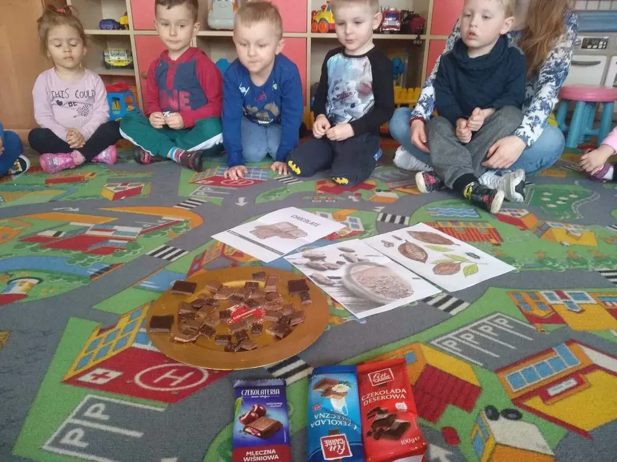 Dzieci w przedszkolu siedzą na dywanie z zabawkami, przed nimi talerz z kawałkami czekolady i opakowania czekolad.