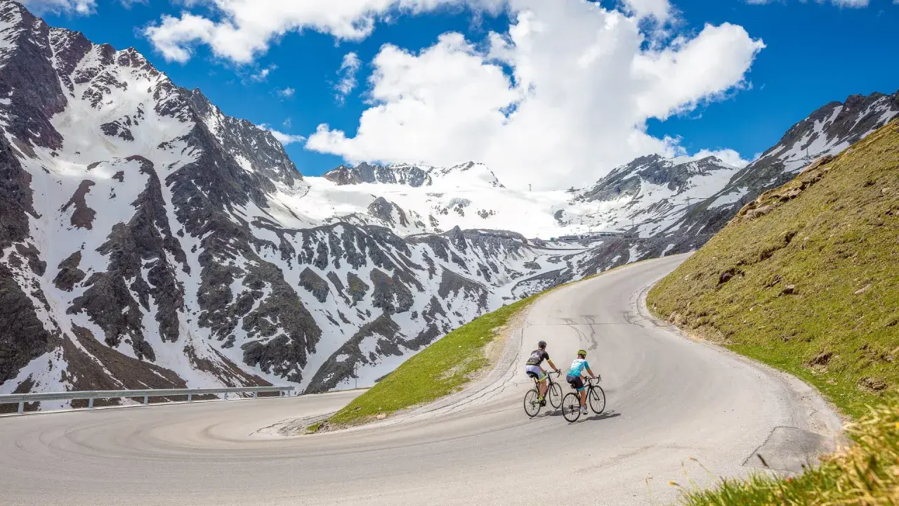 Two cyclists enjoy an epic adventure on a winding road, part of amazing austria bike tours, with snow-capped mountains in the background.