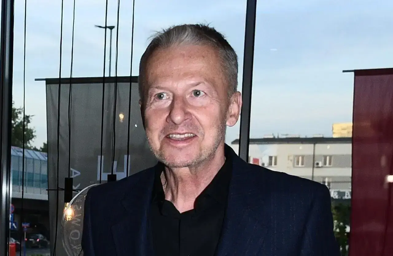 Bogusław Linda w granatowej marynarce i czarnej koszuli, uśmiecha się.