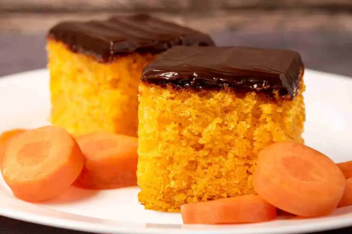 Due fette di torta di carote, soffice e arancione, ricoperte da glassa al cioccolato fondente. Accanto, fette di carota fresca.