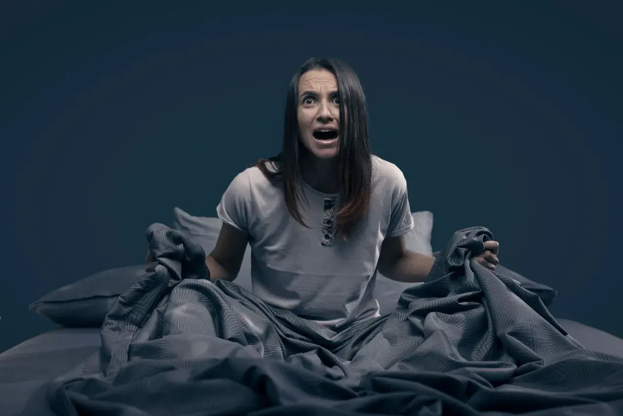 Mujer en la cama, con expresión de angustia, sujetando las sábanas. Parece que se despierta sobresaltada porque me encuentro mal por las mañanas.