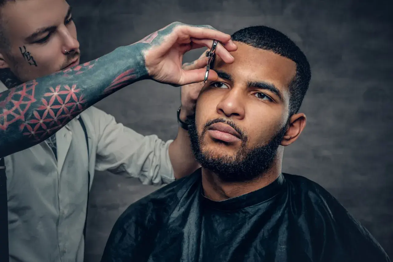 Barbero con tatuajes en el brazo recorta las cejas a un hombre con un corte buzz cut y barba.