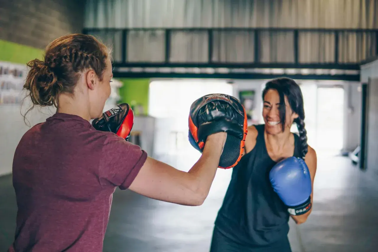 Due donne si allenano nel **boxing** in palestra. Una tiene i guantoni da allenamento, l'altra indossa un guantone blu.