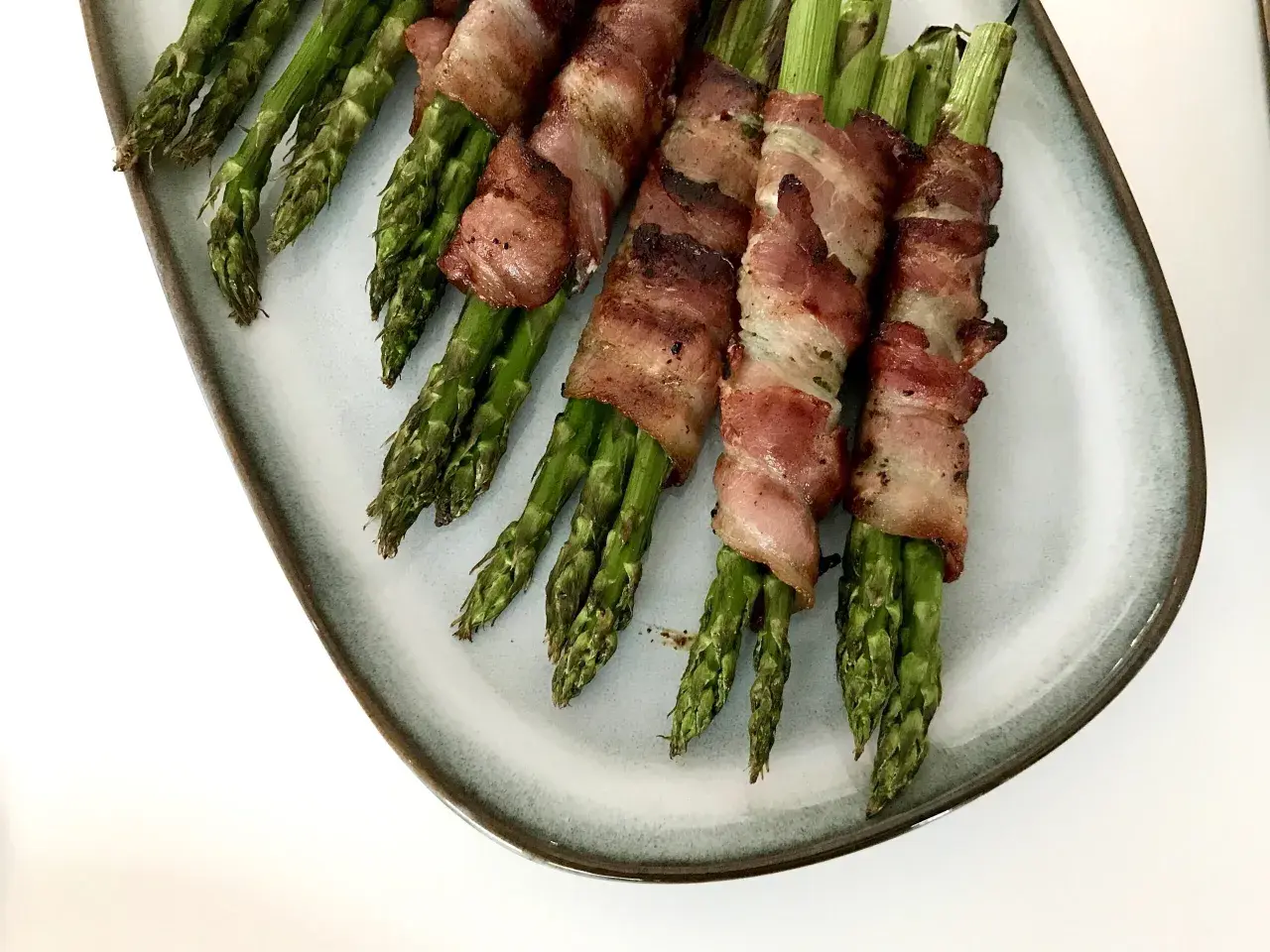 Grön sparris insvept i knaprigt bacon, serverad på en ljusblå tallrik. En utsökt kombination av grönsaker och baconlindad sparris.