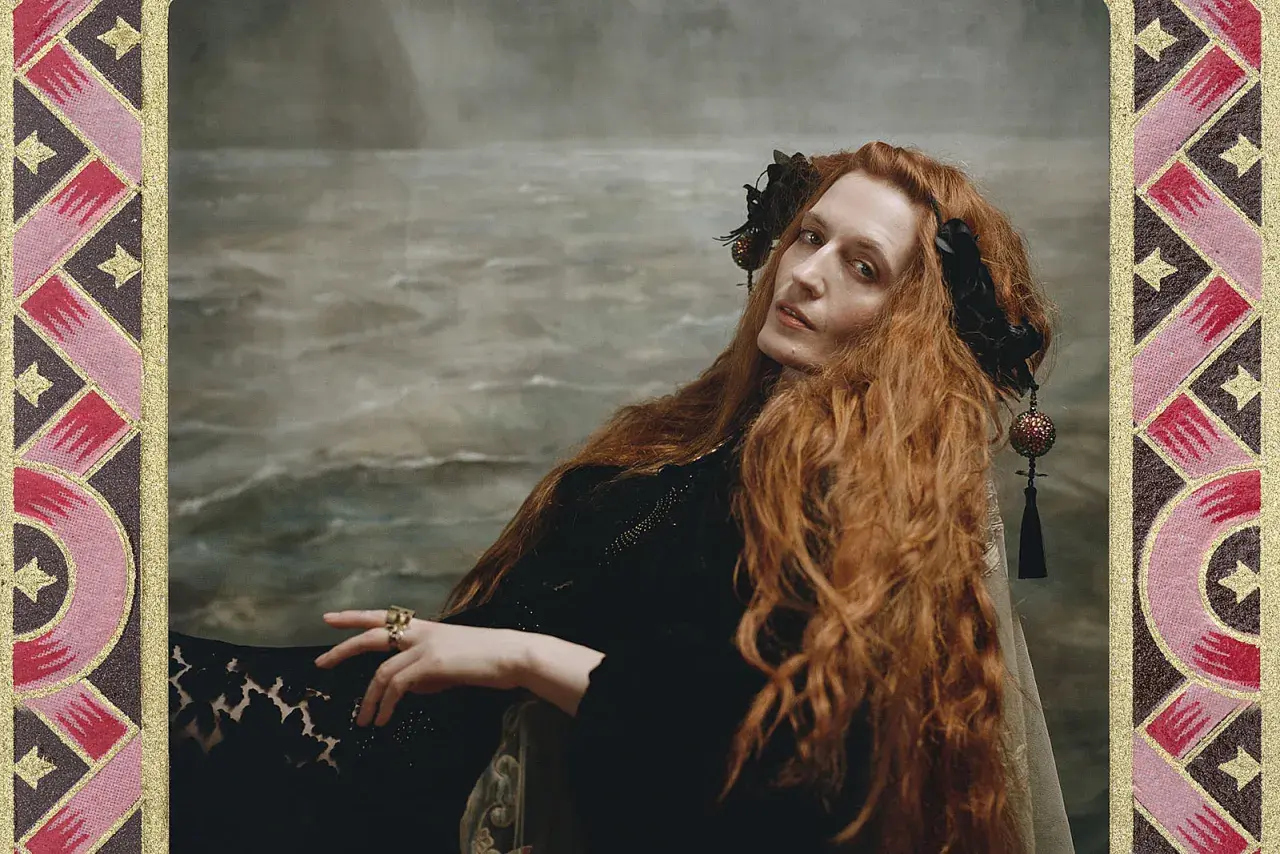 Florence Welch, jak królowa, z rudymi włosami i w czarnej sukni, na tle morskiego krajobrazu.