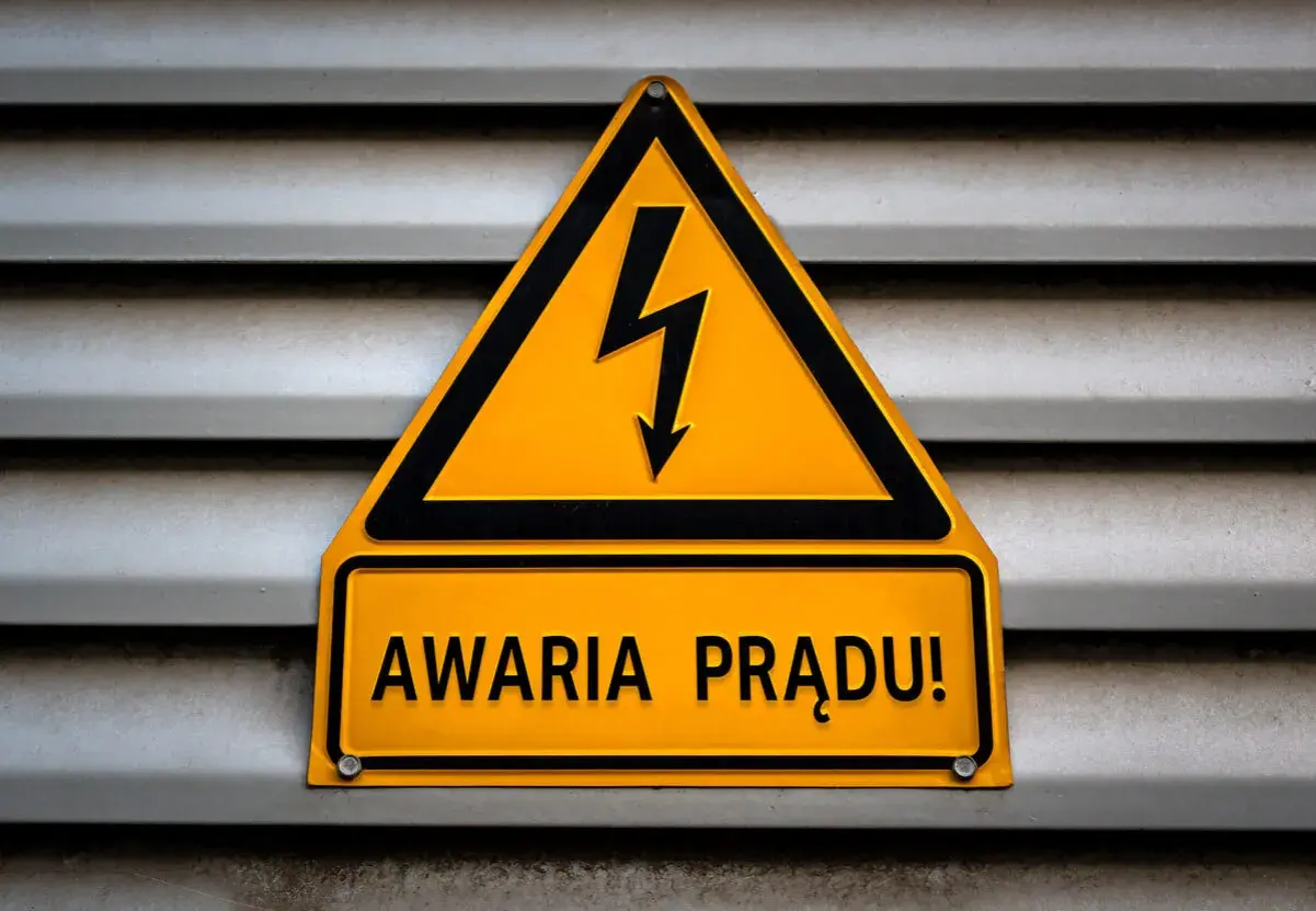 Żółty trójkąt z symbolem pioruna i tabliczka "AWARIA PRĄDU!" na szarej, metalowej powierzchni.