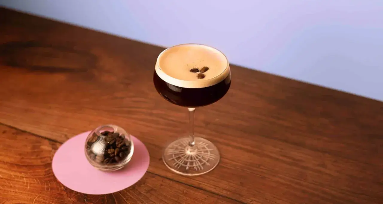 Espresso Martini with coffee beans on top, next to a small glass of coffee beans on a pink coaster.