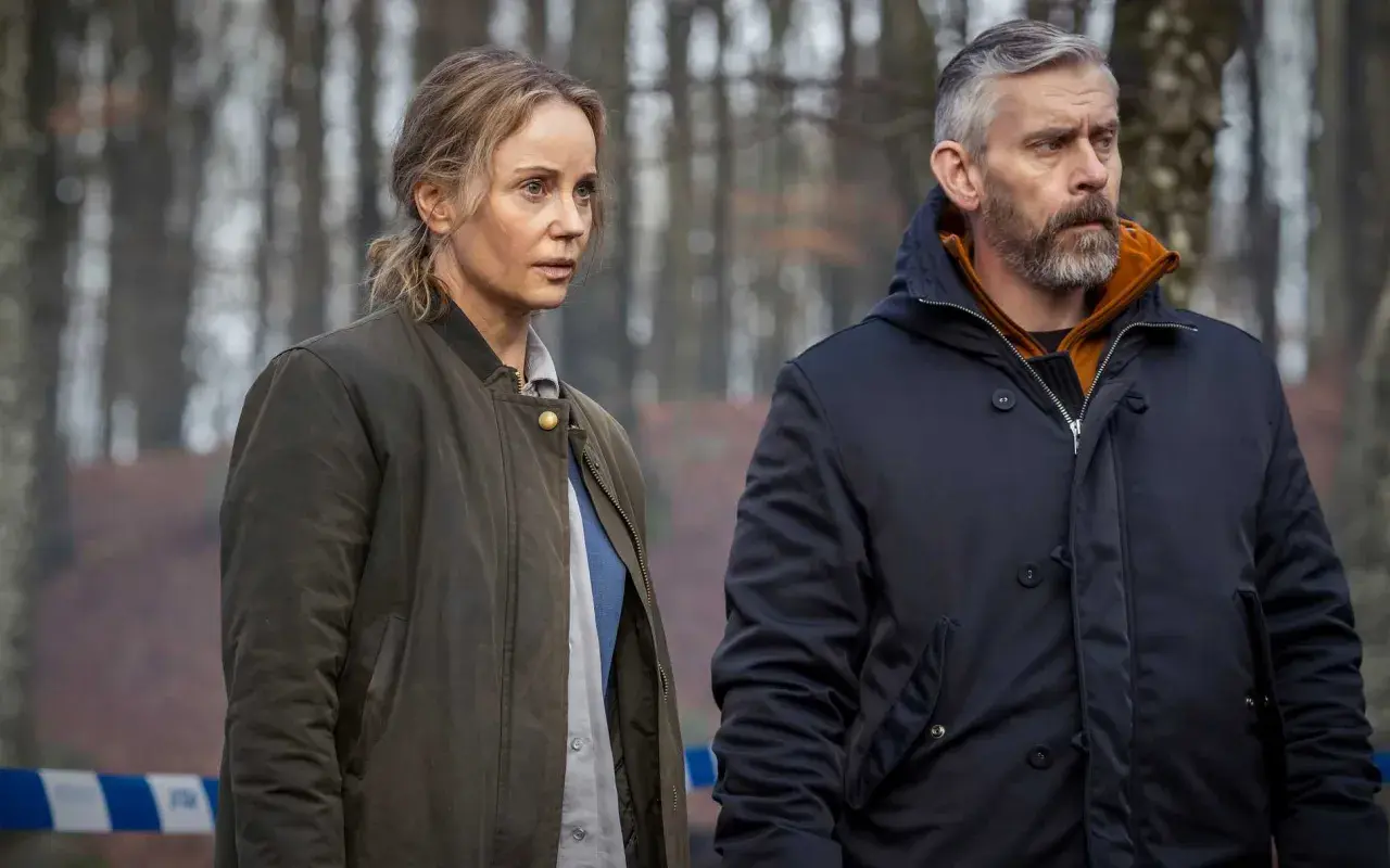 Två personer, en kvinna och en man, står i en skog. Bakom dem syns blåvit avspärrningsband. Kanske är det Sandor Berg som utreder ett brott.