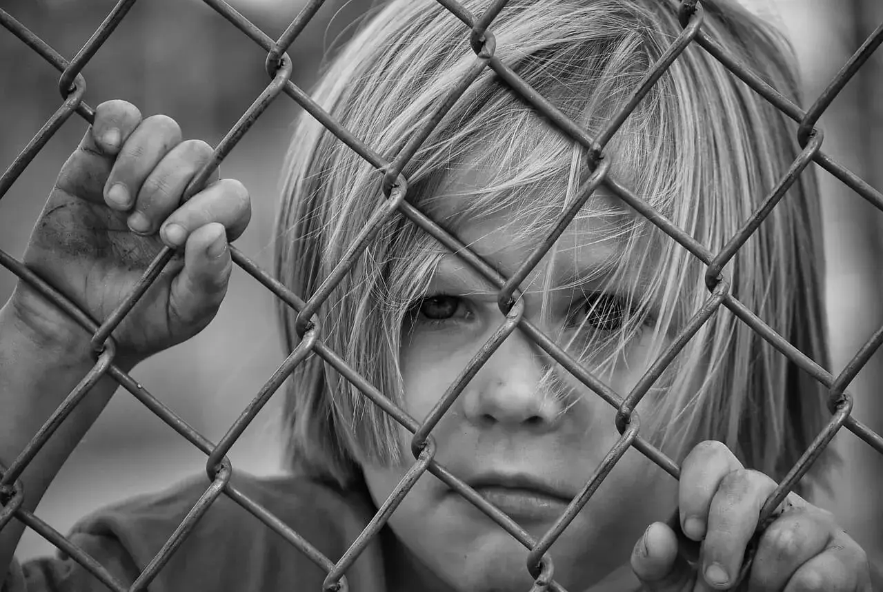 Czarno-białe zdjęcie dziecka z blond włosami, patrzącego przez siatkę ogrodzeniową.