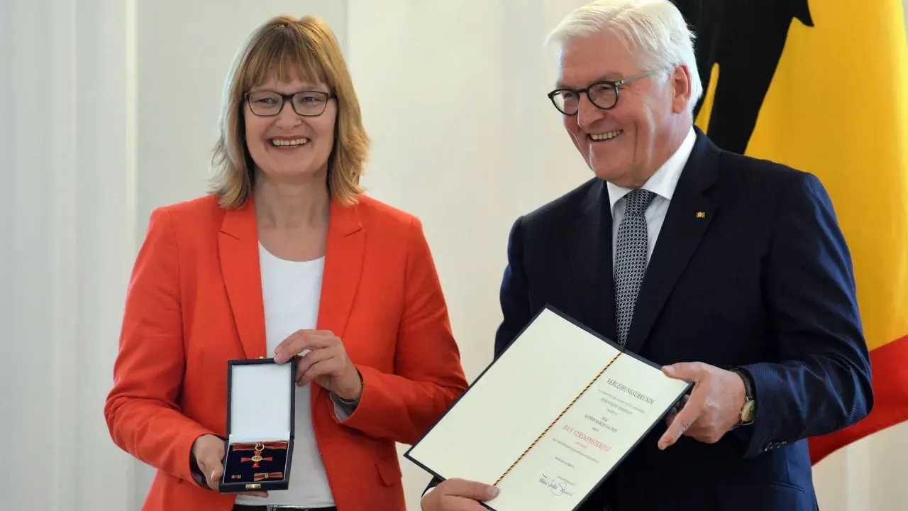 Frau mit rotem Blazer erhält das Bundesverdienstkreuz. Präsident Steinmeier überreicht die Auszeichnung und Urkunde.