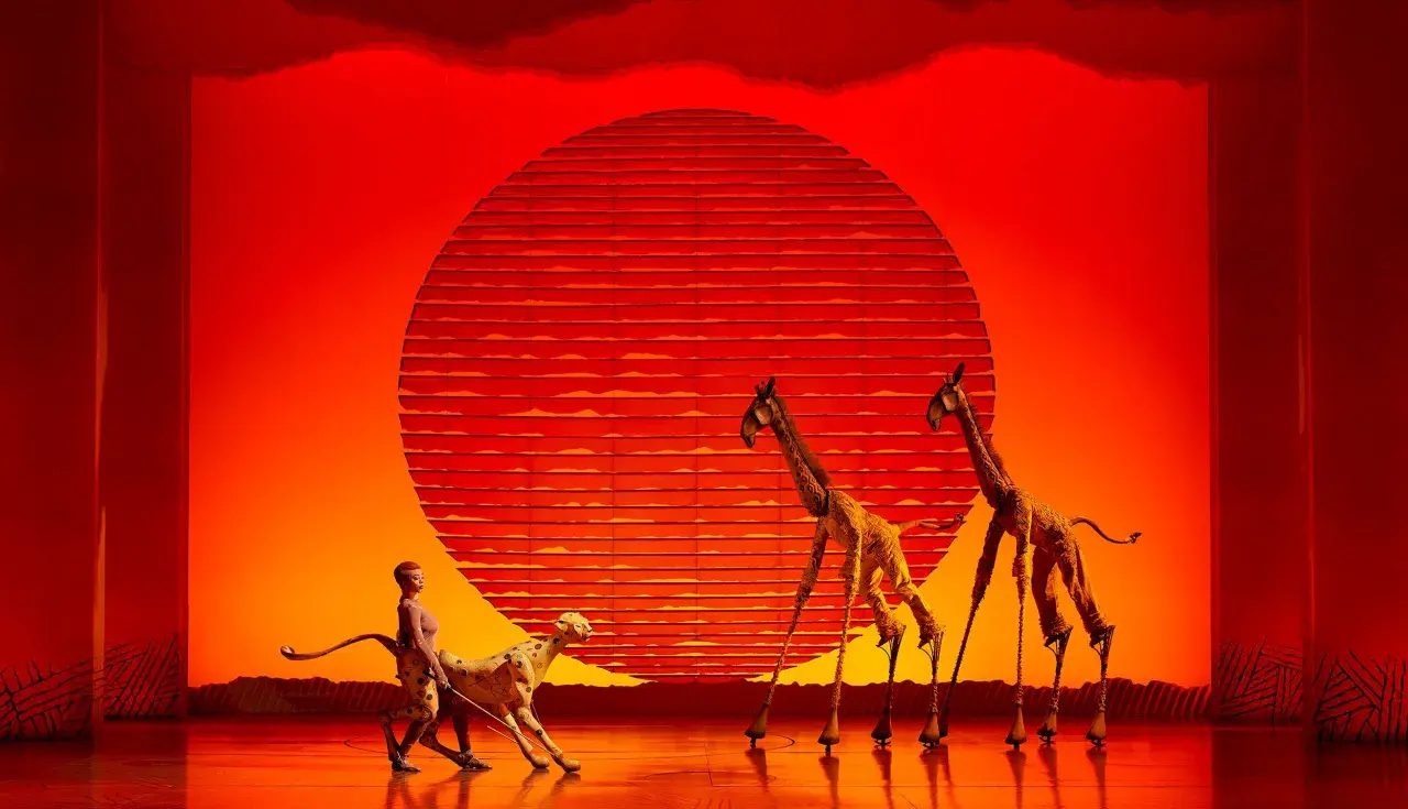 Escena de musicales en Zaragoza: un guepardo y dos jirafas en un escenario con un sol rojo.