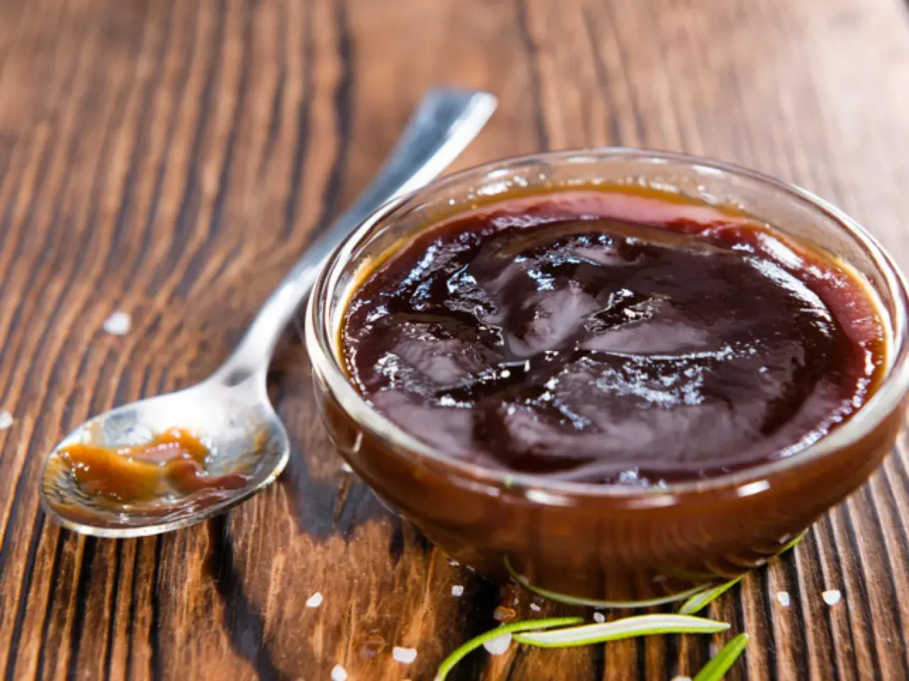 Un cuenco de cristal lleno de deliciosa salsa barbacoa casera, con una cuchara al lado y romero fresco.