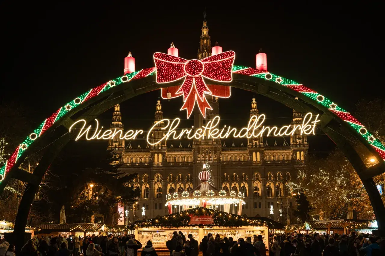 Un mercado navideño en Viena, con un arco de luces y un gran lazo rojo. El edificio del ayuntamiento ilumina el fondo.