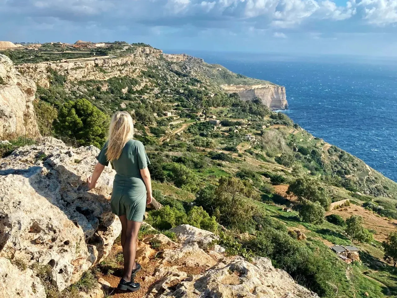 Malta, luty. Pogoda słoneczna, wietrzna. Kobieta na klifie podziwia widok na morze i zielone wzgórza.