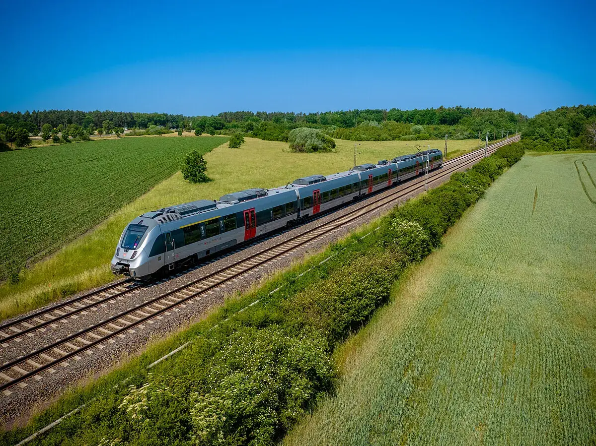 Moderner Triebzug fährt auf einer **railway deutsch** durch grüne Felder und Wälder unter blauem Himmel.