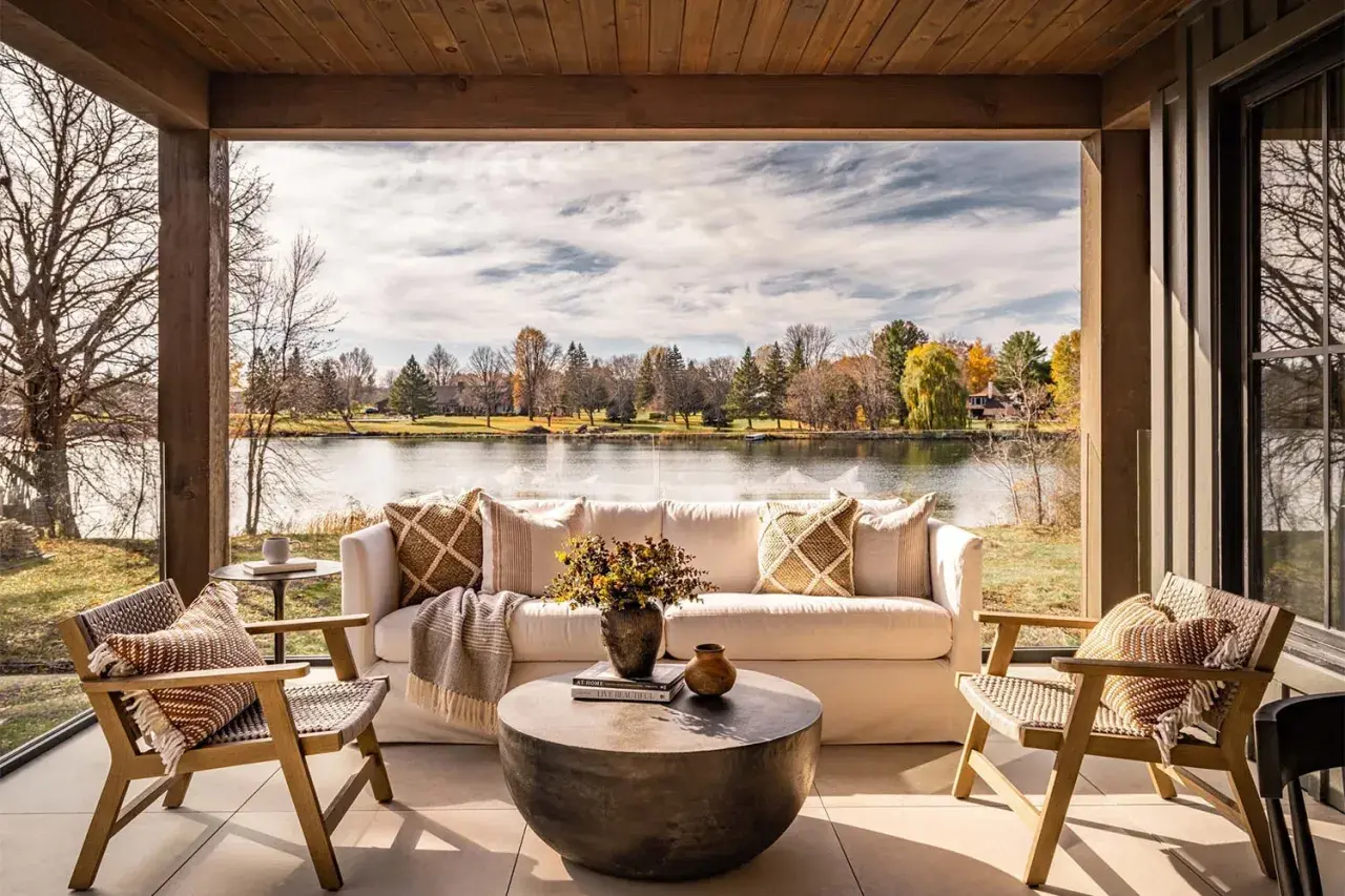 Cozy lakehouse interior design with a white sofa, chairs, and coffee table on a covered patio overlooking a serene lake and autumn foliage.