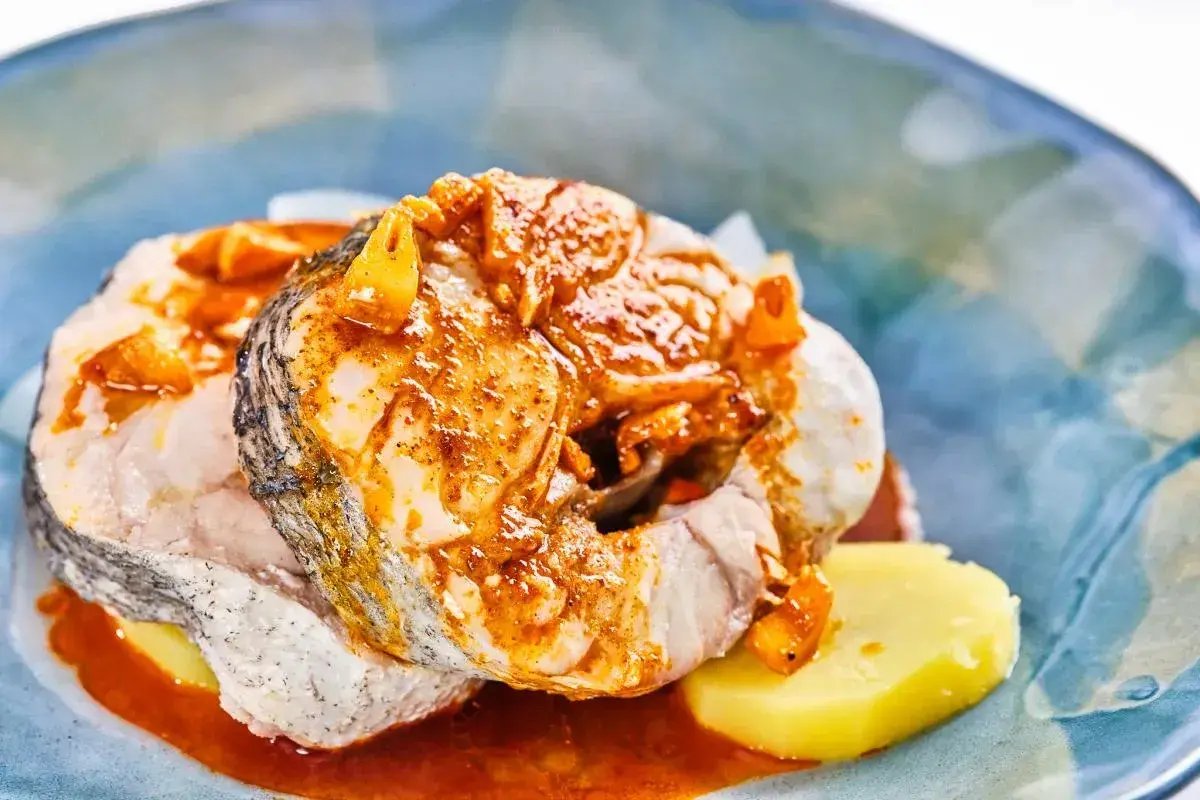 Rodajas de pescado blanco cubiertas con una sabrosa salsa gallega, servidas sobre patatas cocidas en un plato azul.