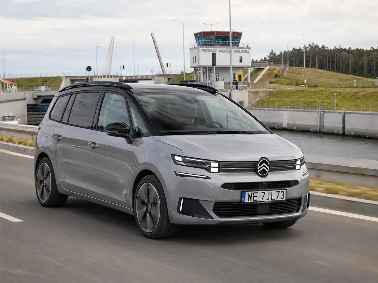 Srebrny Citroen Picasso jedzie drogą obok kanału. W tle widoczna wieża kontrolna i podniesiony most.