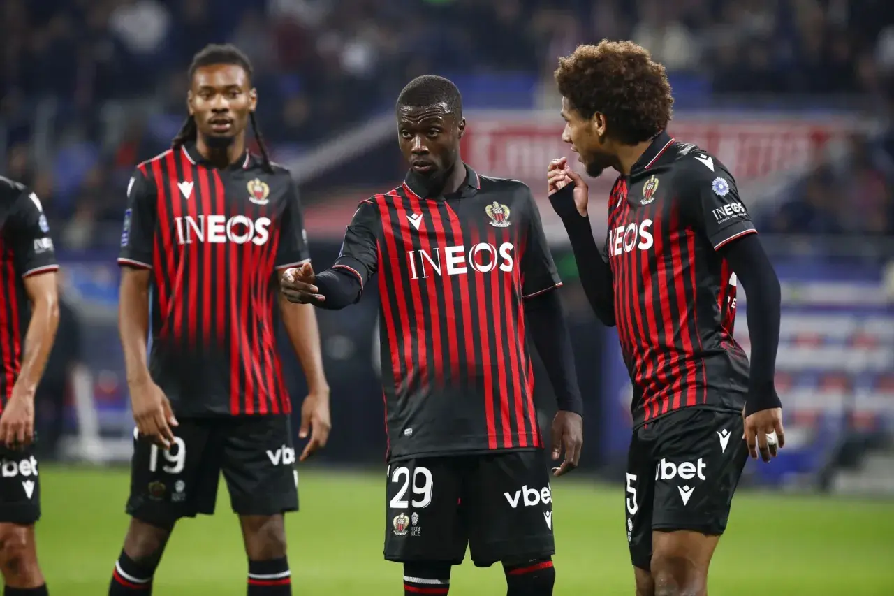Piłkarze OGC Nice w czarno-czerwonych strojach dyskutują na boisku. Jeden z nich, z numerem 29, wskazuje palcem.