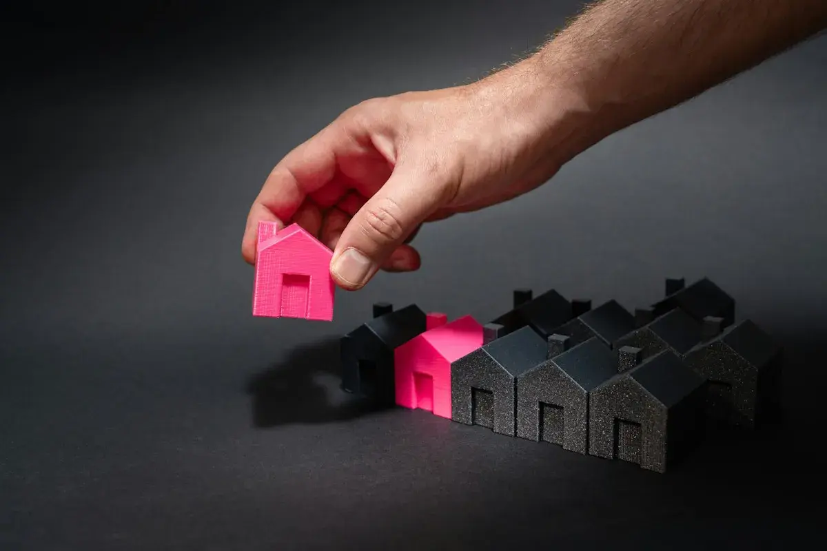 Mano che sceglie una casetta rosa tra tante nere. Un'idea di permuta immobiliare con costruttore.