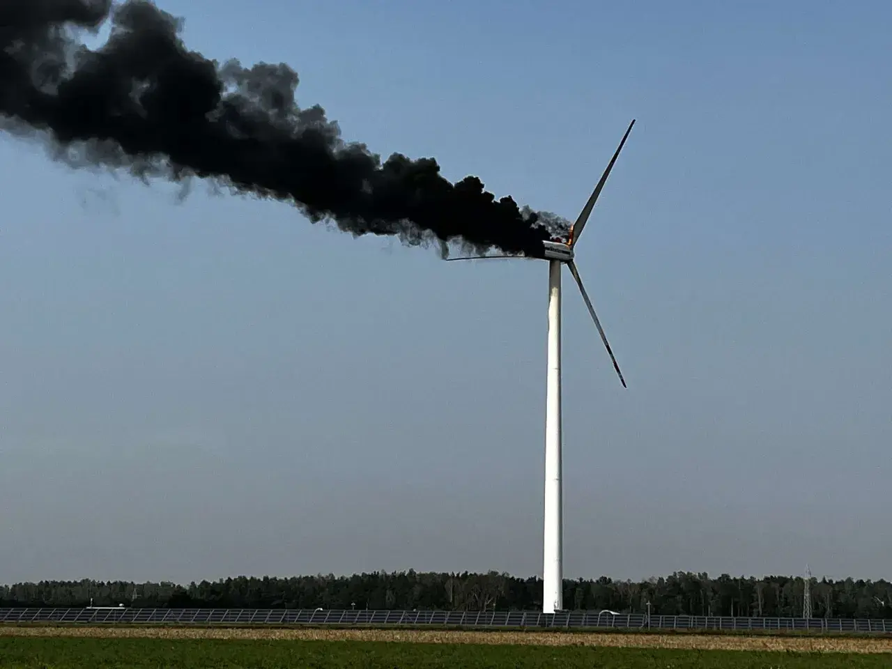 Płonąca turbina wiatrowa z gęstym, czarnym dymem unoszącym się na tle błękitnego nieba.