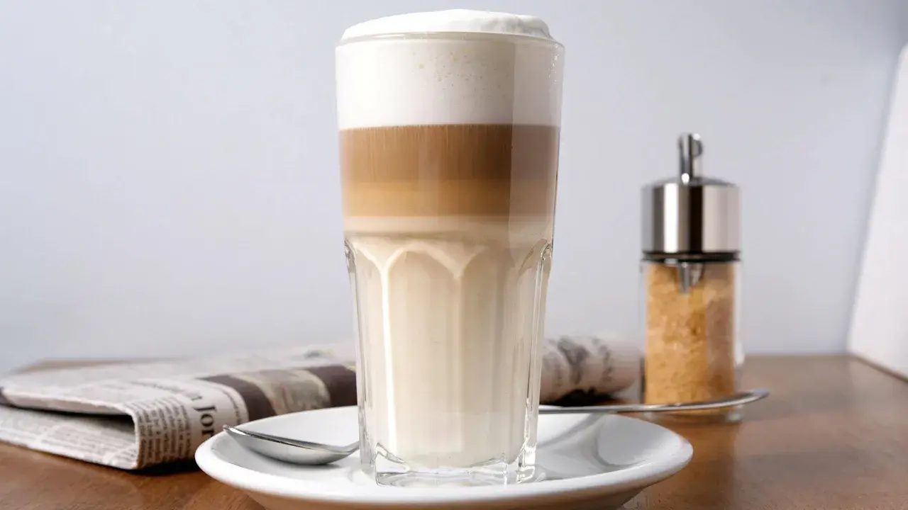 Ein geschichteter Latte Macchiato im Glas, daneben eine Zeitung und ein Zuckerspender. Milchkaffee vs. Latte Macchiato – die Wahl ist schwer!