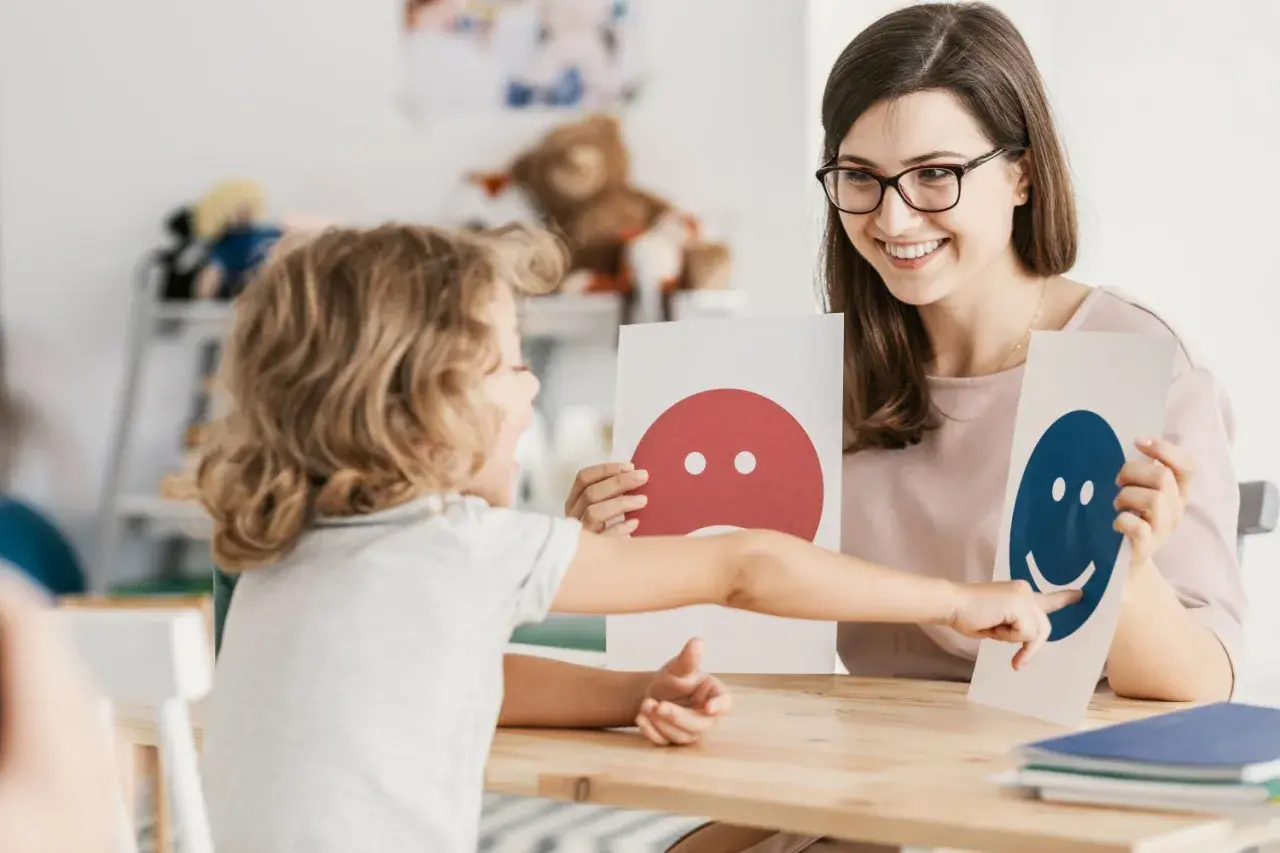 Dziecko wskazuje na uśmiechniętą buźkę, rozwijając inteligencję intrapersonalną. Terapeuta z uśmiechem prezentuje karty emocji.