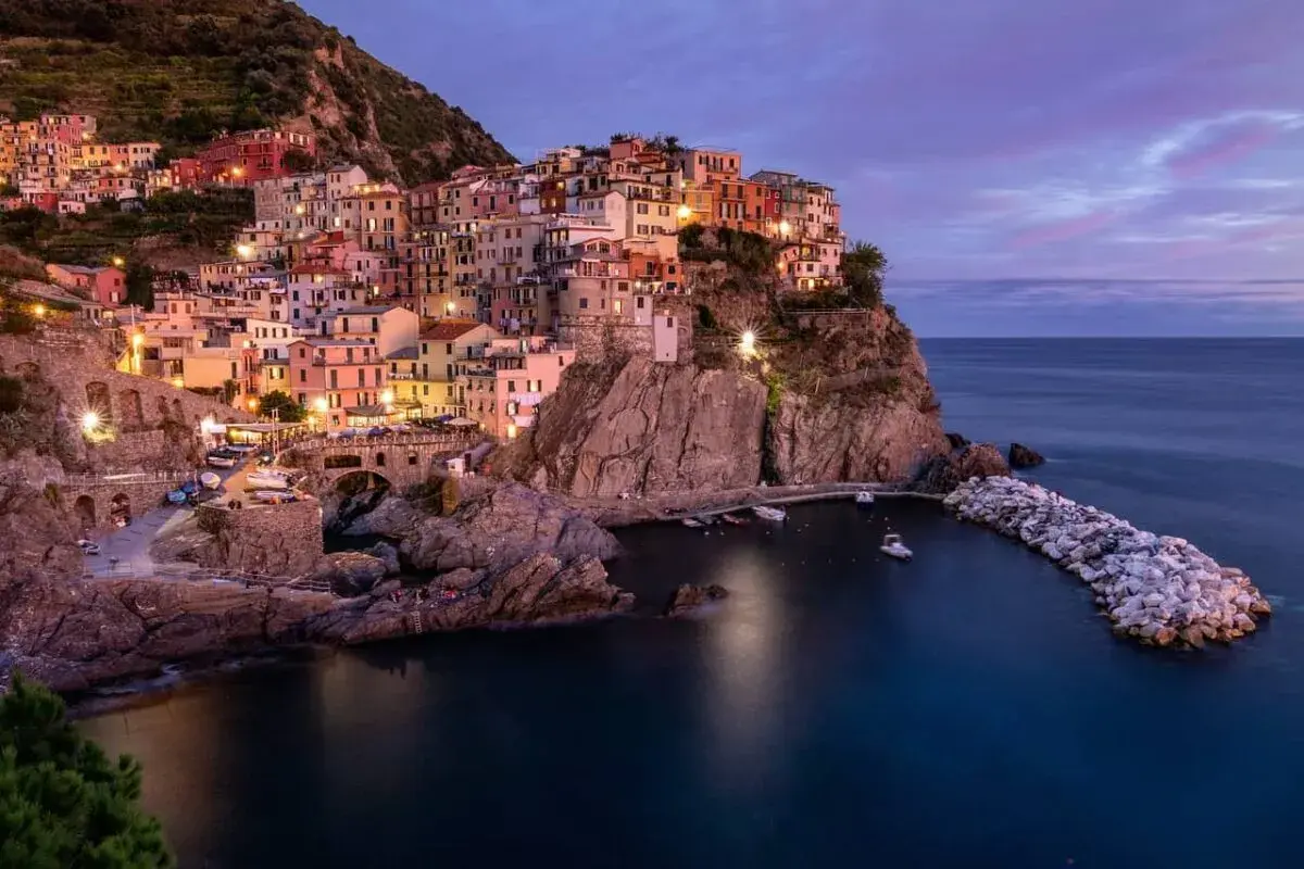 Manarola, una delle perle della strada panoramica 5 Terre, si illumina al crepuscolo. Case colorate aggrappate alla roccia, un piccolo porto e il mare blu intenso.
