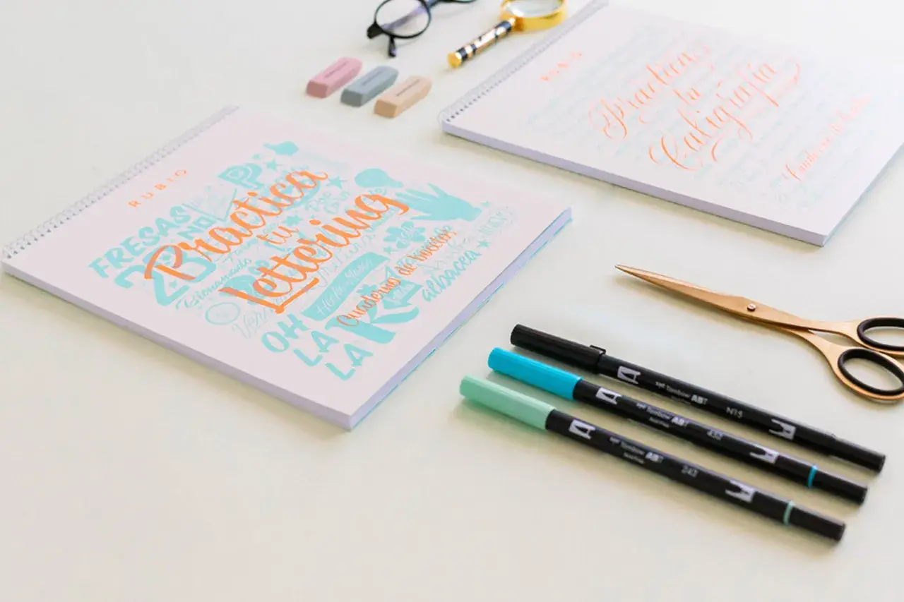 Cuadernos de "Practica Lettering" y "Practica tu Caligrafía" con rotuladores y tijeras doradas.