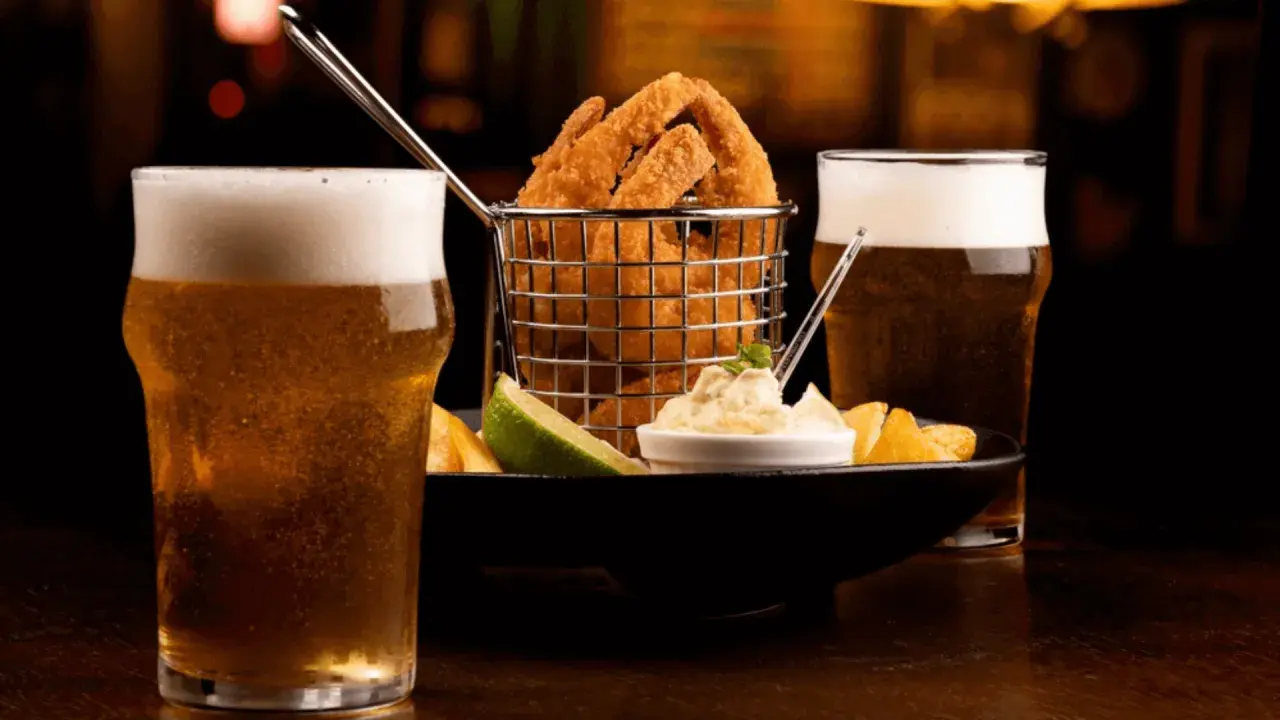 Due bicchieri di birra bionda ghiacciata accompagnano un cestino di anelli di cipolla fritti, patatine e una salsa cremosa.
