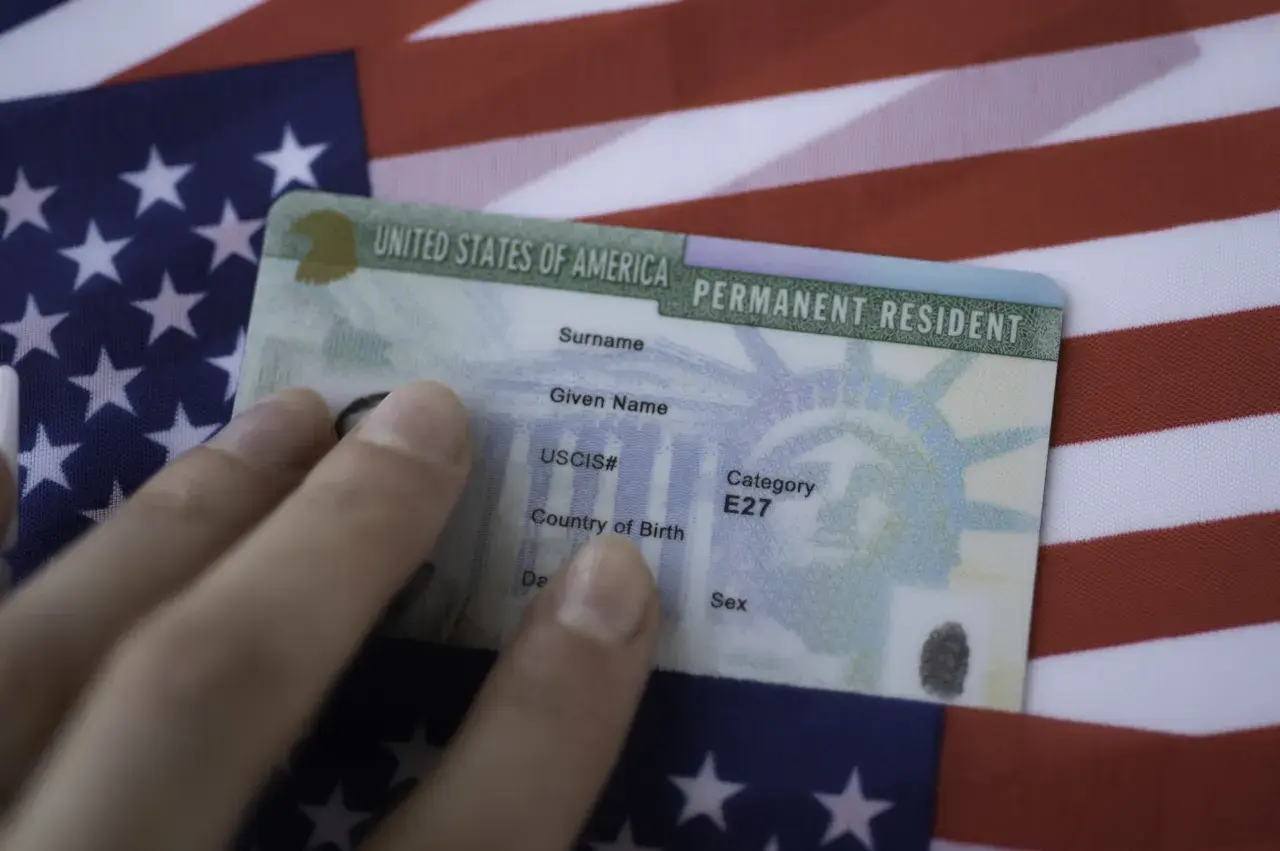 Dłoń trzyma zieloną kartę, dokument potwierdzający status stałego rezydenta USA, na tle amerykańskiej flagi. To dowód na to, co to zielona karta.